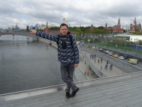 На Парящем (над рекой Москва) мосту с видом на Кремль, оперевшись рукой на стеклянное ограждение, которого почти не видно.